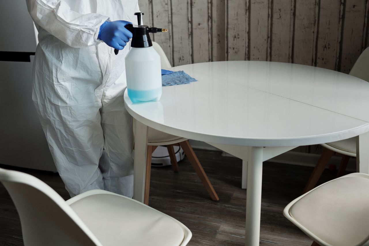 team-04 A person in a protective suit sanitizing a white table indoors with a spray bottle.